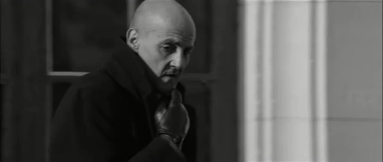 13 Tzameti (2006) movie still: medium — A man stands inside a building near a windowed corridor with high contrast lighting. The man wears a dark…