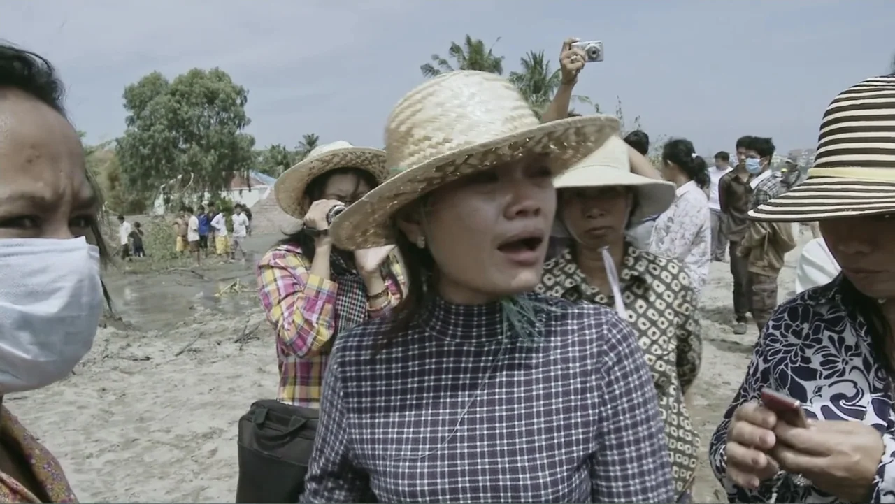 A Cambodian Spring (2018) movie still: medium — A group of people gathers on a sandy riverbank during the day. Multiple individuals wear hats and…