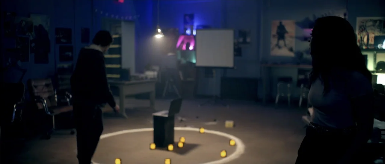 Elevator Game (2023) movie still: wide — A dark, cluttered basement room contains a white circle painted on the concrete floor. Two people stand in…