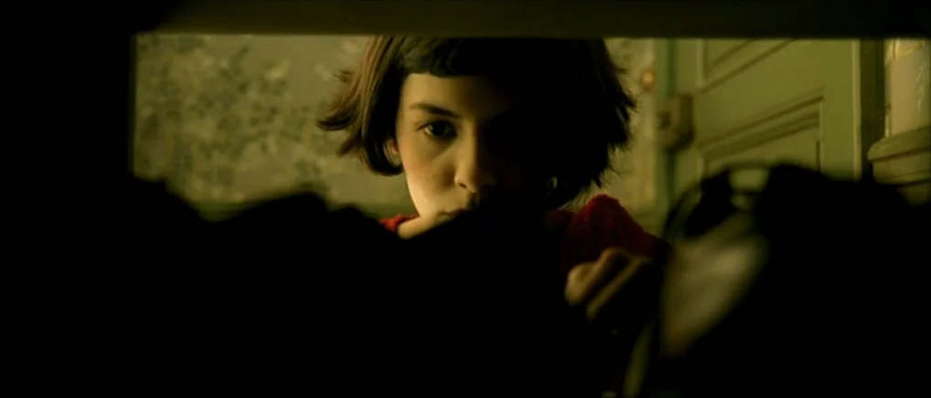 Amelie (2001) movie still: closeup — A woman looks through a dark opening into a concealed, dimly lit shelf space. The background features…