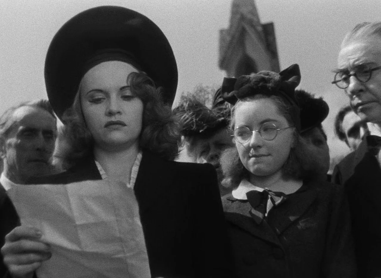 Le Corbeau (1943) movie still: medium — A group of people gathers outdoors in front of a stone gothic structure. A woman holds a piece of paper and…