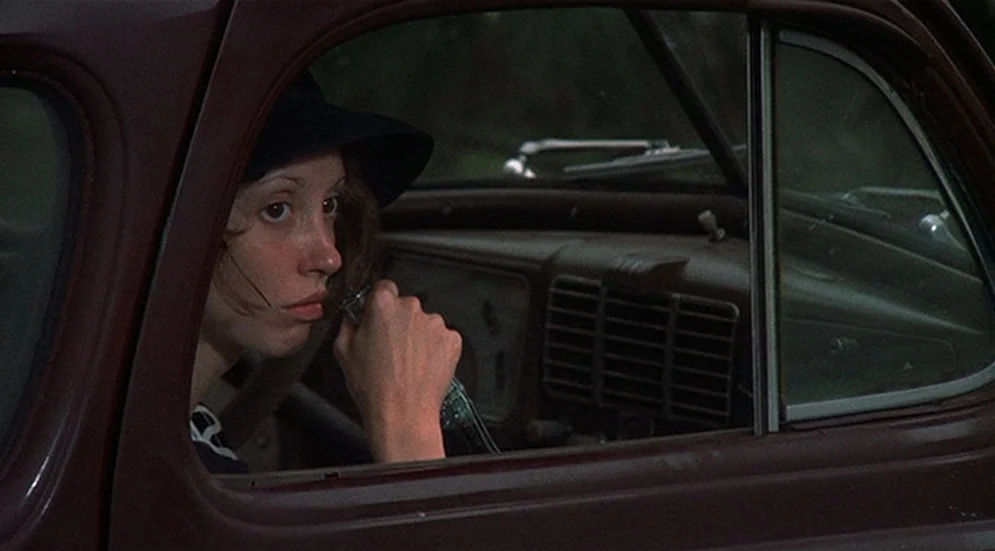 Thieves Like Us (1974) movie still: medium — A vintage maroon car interior shows a woman sitting in the driver seat. She holds a glass bottle in her…