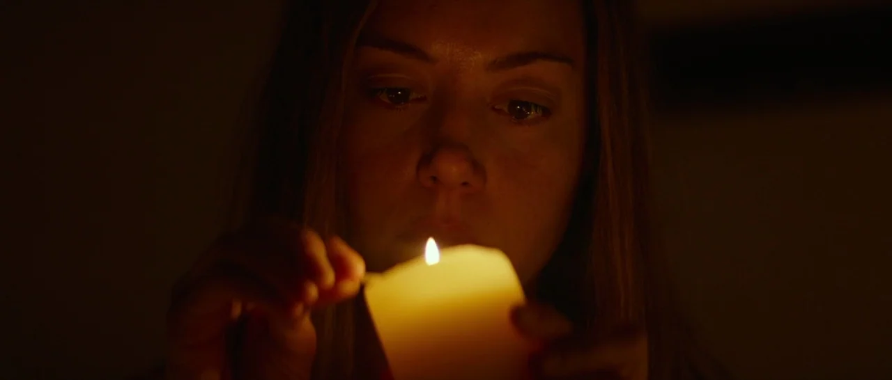 Ingrid Goes West (2017) movie still: closeup — A dark, undetailed interior environment features a person lit by candlelight. The subject holds a…