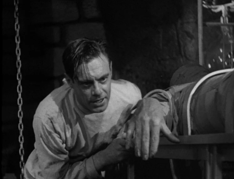 Frankenstein movie still: medium — A stone-walled laboratory contains a metal operating table with a figure strapped down. A man in medical clothing…