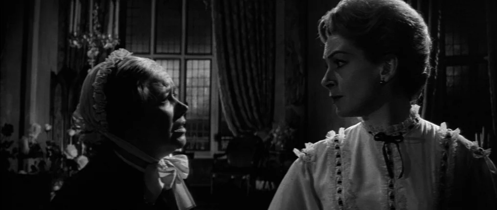 The Innocents (1961) movie still: medium — An opulent, dimly lit room with large windows and heavy curtains in the background. Two women converse…