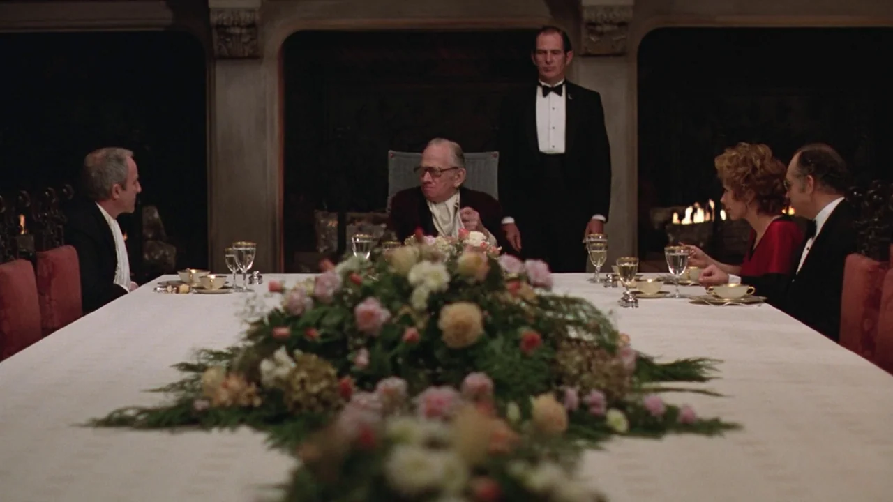 Being There (1979) movie still: wide — A formal dining room features a long table with a central floral arrangement. Five people participate in a…