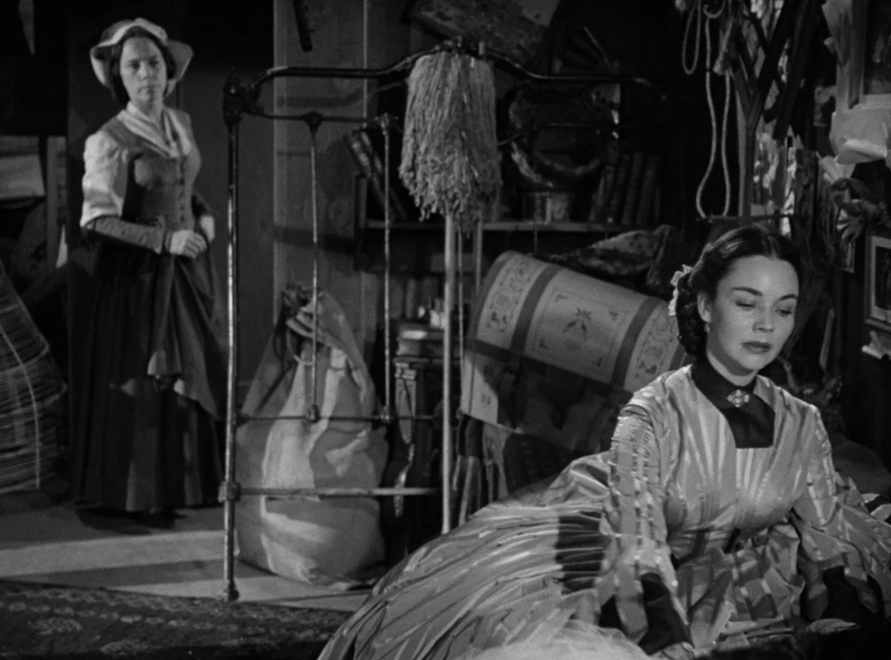 Madame Bovary (1949) movie still: medium — An attic room filled with various household items, trunks, and draped fabric. One woman lies down in the…