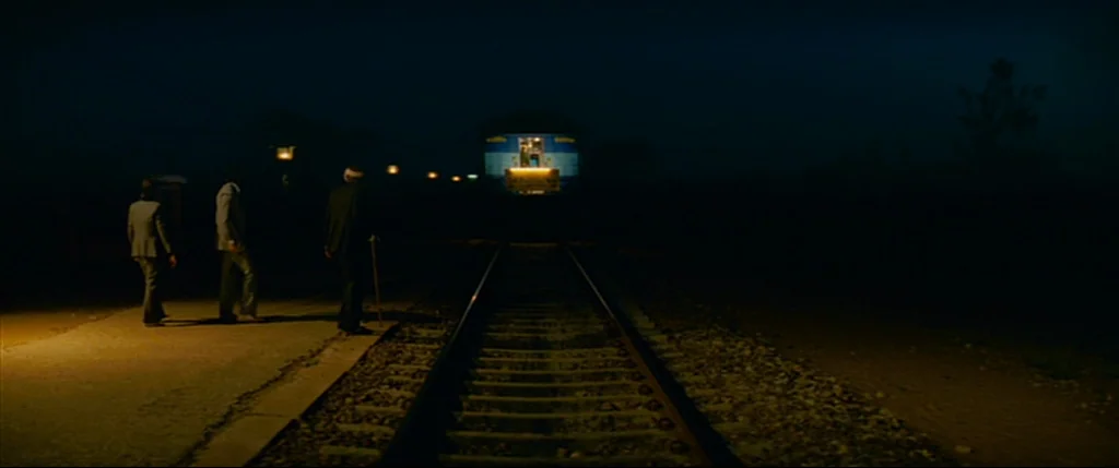 The Darjeeling Limited (2007) movie still: wide — A desolate train platform at night features three men standing near the tracks. The men walk…