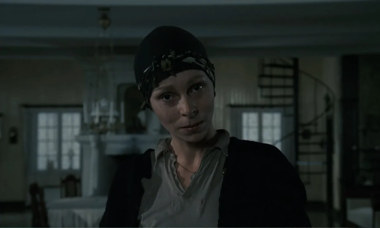 The Sacrifice (2015) movie still: medium — A dimly lit room with a spiral staircase visible in the background shadow. A woman wearing a black head…