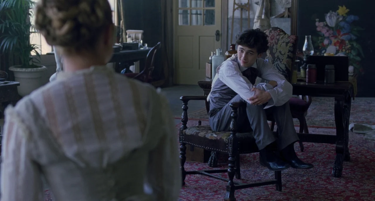 Little Women (2019) movie still: medium — An artist studio filled with antique chairs, paintings, and jars on wooden tables. A young man sits in a…