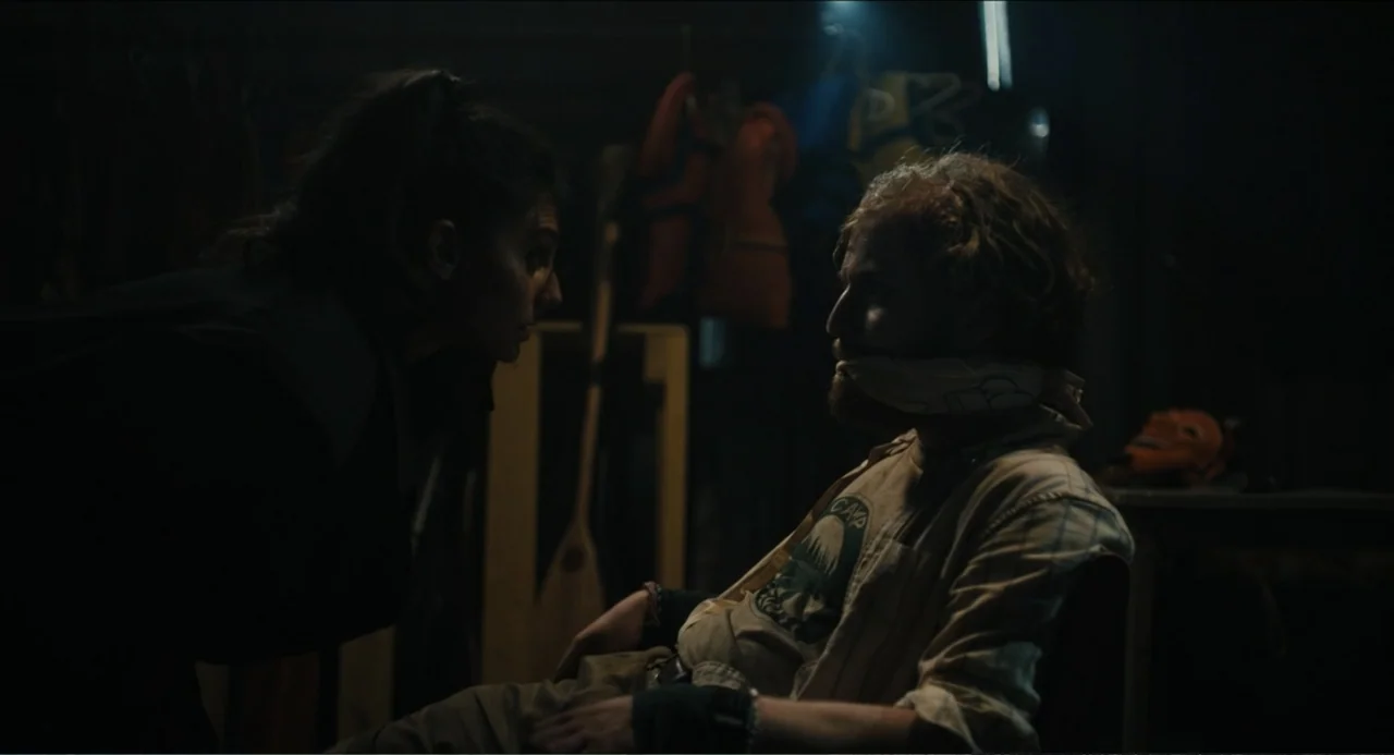 Hell of a Summer (2025) movie still: medium — A dark indoor storage space contains scattered gear and hanging equipment in the background. A woman…