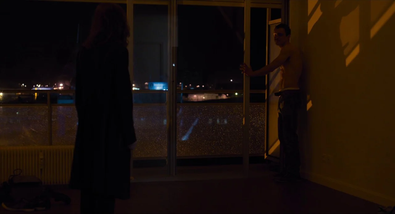 Happy End (2017) movie still: wide — An interior space faces an open balcony at night with city lights in the distance. A woman stands in the…