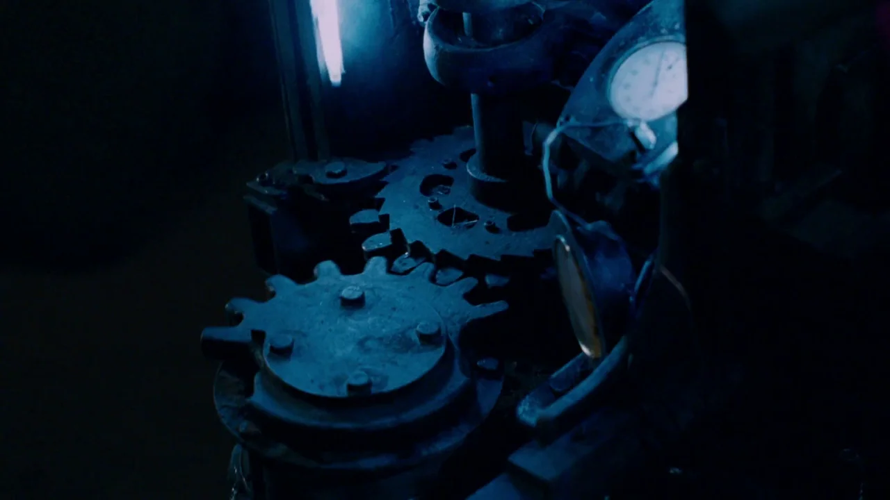 Saw IV movie still: closeup — Industrial metal gears are positioned within a dark machine housing. A pressure gauge is visible on the right side of…