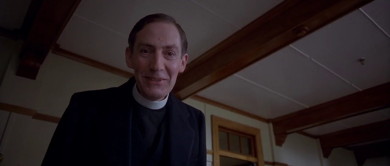 Heavenly Creatures (1994) movie still: medium — A low-angle view shows a priest standing within a plain, wooden-beamed building interior. The man…