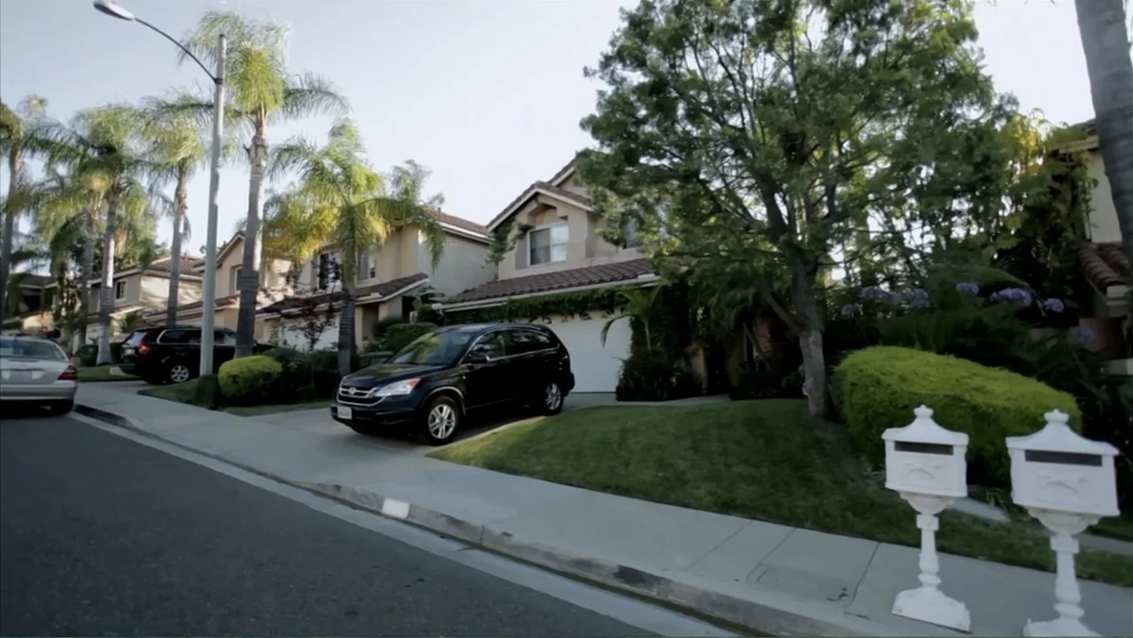 The (Dead Mothers) Club (2014) movie still: wide — A quiet suburban street features rows of houses with manicured lawns and palm trees. Two white…