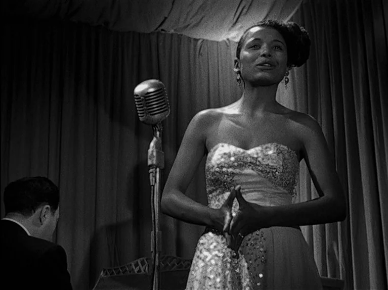 The Big Night (1951) movie still: medium — A dimly lit lounge or nightclub with heavy curtains hanging in the background. A woman in a sequined gown…