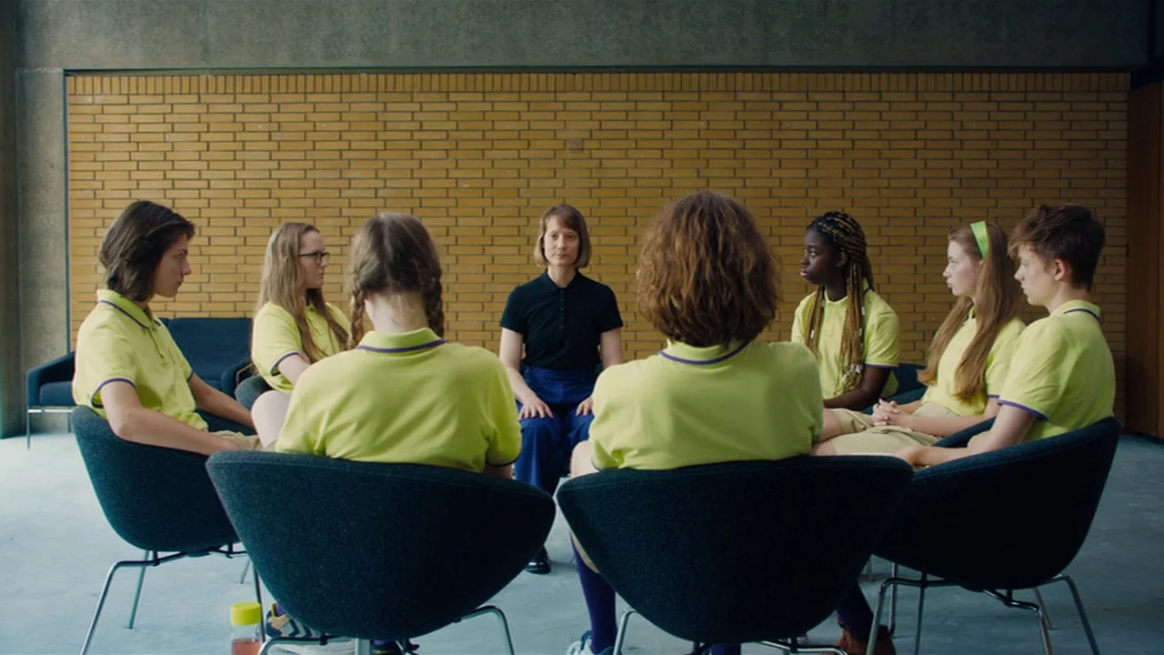 Club Zero (2023) movie still: wide — A group of teenagers and one adult sit in a circle in a room. The room has a textured yellow brick wall and…