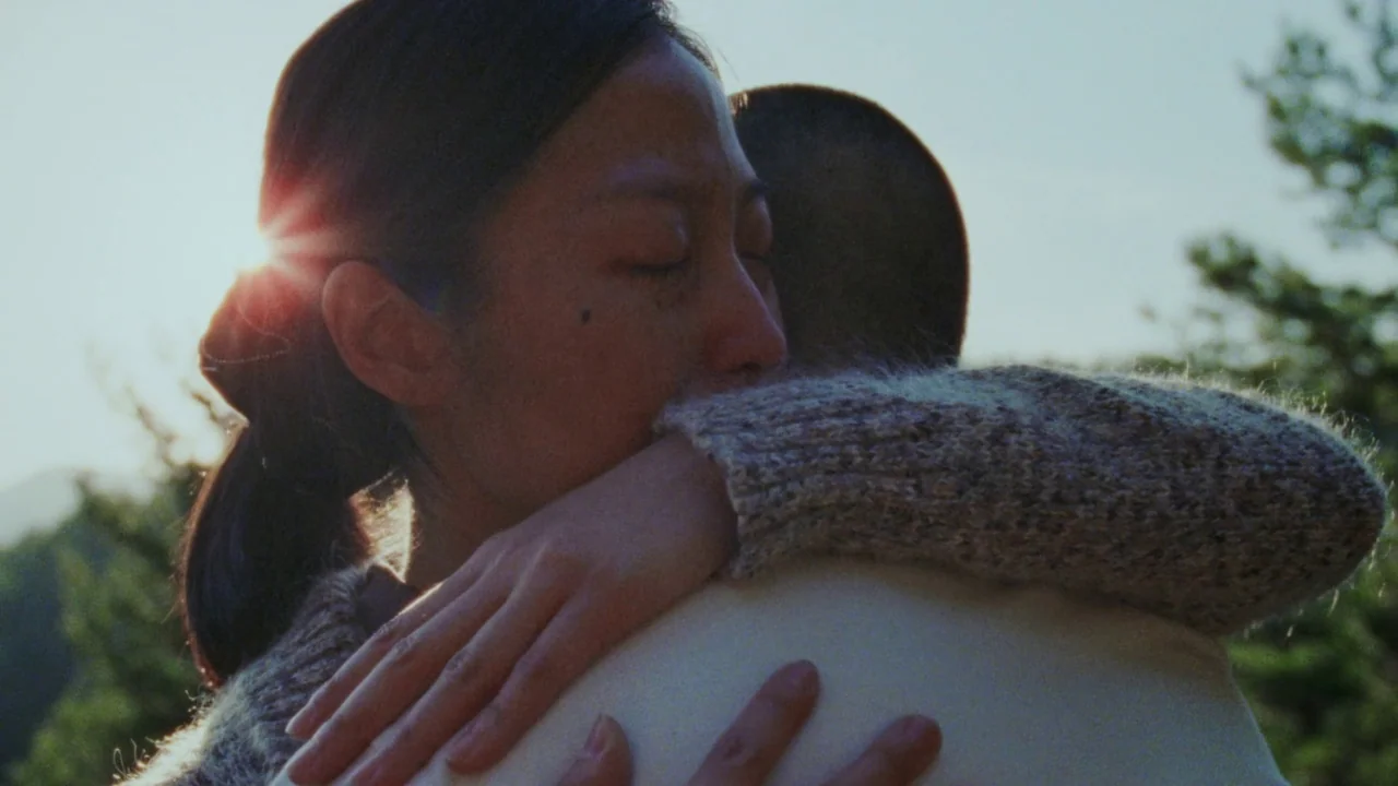 Riceboy Sleeps (2022) movie still: closeup — Two people embrace in an outdoor setting with a bright sun background. Sunlight flares behind the…
