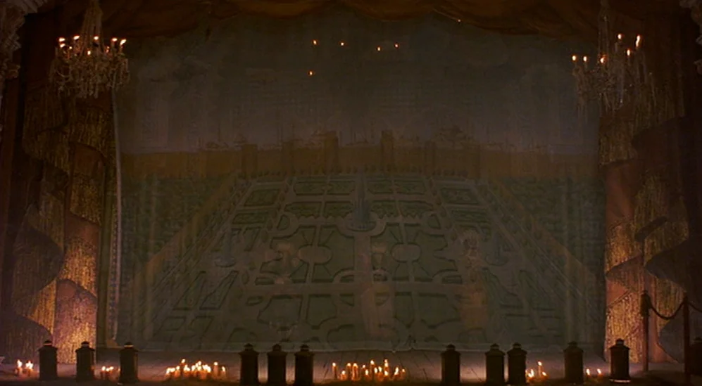 The Adventures Of Baron Munchausen (1988) movie still: establishing — A dimly lit theater stage features a large painted backdrop showing a…