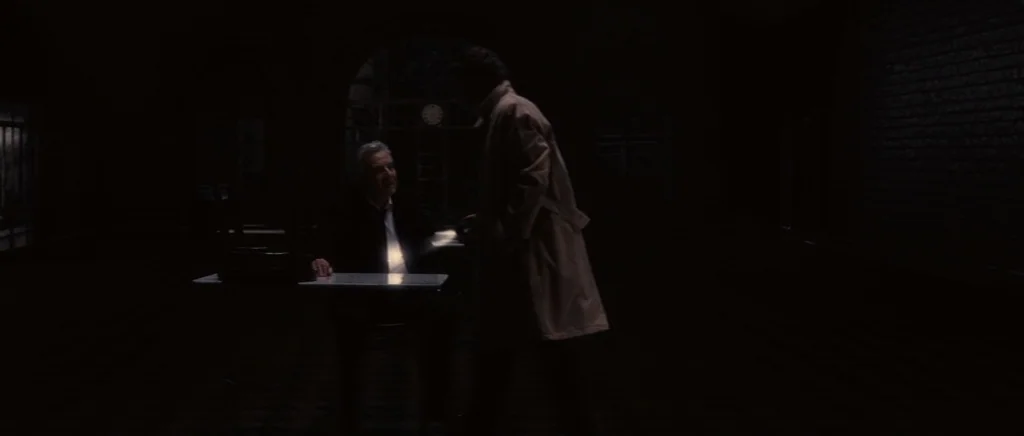You Ain’t Seen Nothin’ Yet (2012) movie still: wide — A dark, sparsely furnished room with a single desk illuminated by an overhead light. A man…