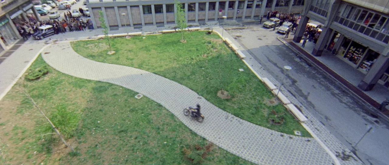 What Have They Done To Your Daughters? (1974) movie still: establishing — A paved path snakes through a grassy urban plaza surrounded by multi-story…