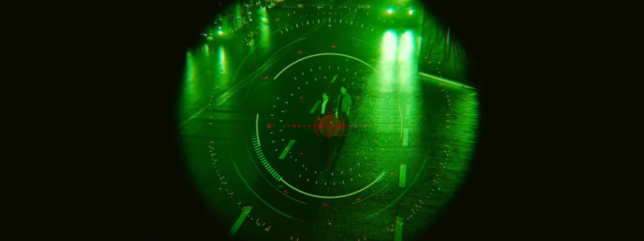 Under The Light (2023) movie still: wide — A dark city street at night is viewed through a circular night-vision scope. Two people stand in the…