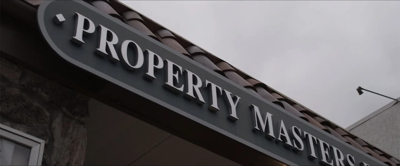 Heavenly Deposit (2019) movie still: closeup — An outdoor commercial storefront entrance features a grey sign with white dimensional text. Brown…