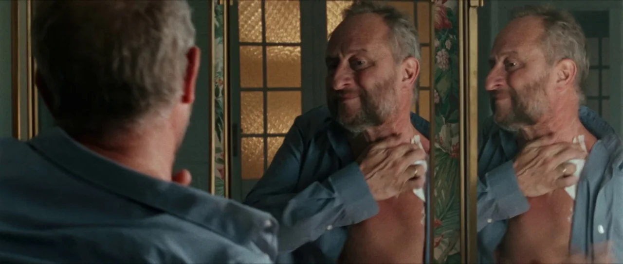 Inexorable (2021) movie still: medium — A vintage-style bathroom features floral patterned wallpaper and a multi-panel mirror frame. An elderly man…