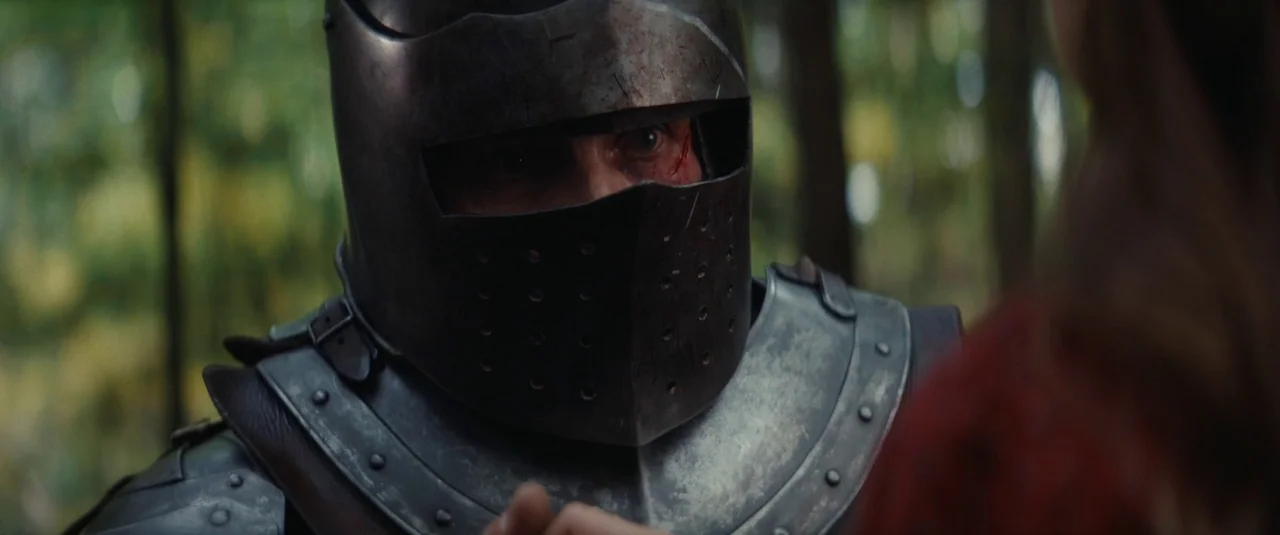 The Dead Don’t Hurt (2023) movie still: closeup — A knight stands in a forest, wearing dark metal armor with a scarred face. He turns his head while…