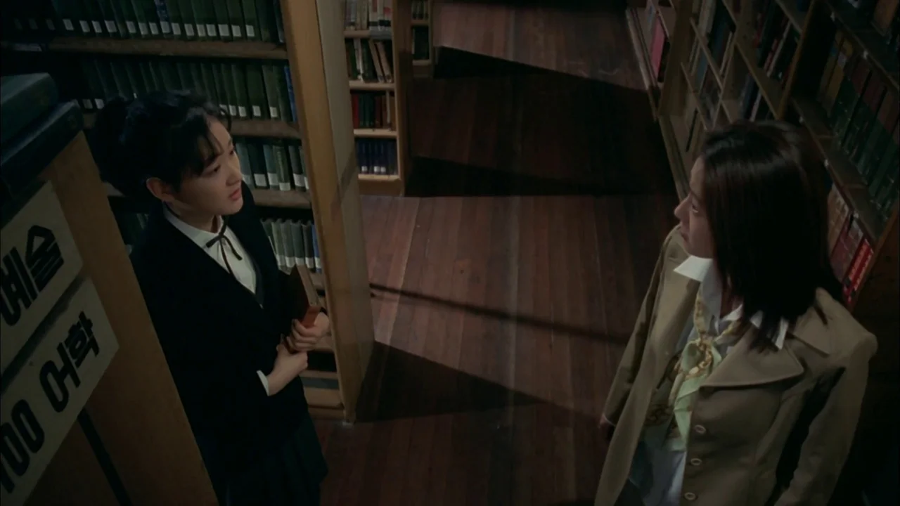 Whispering Corridors (1998) movie still: medium — A narrow library aisle with tall, filled wooden bookshelves and dark polished floorboards. Two…