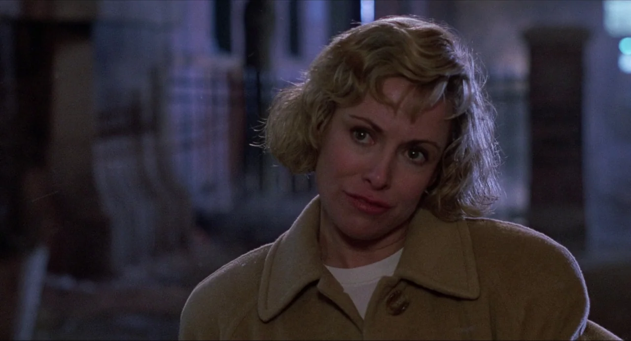 Child’s Play (1988) movie still: closeup — A city street at night with a blurred brick building in the background. A woman in a tan coat looks…