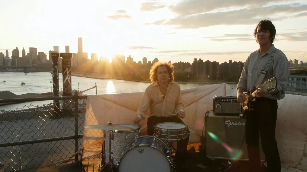 Counting (2015) movie still: wide — An urban rooftop overlooking a river and city skyline during sunset. Two musicians perform with a drum set and…