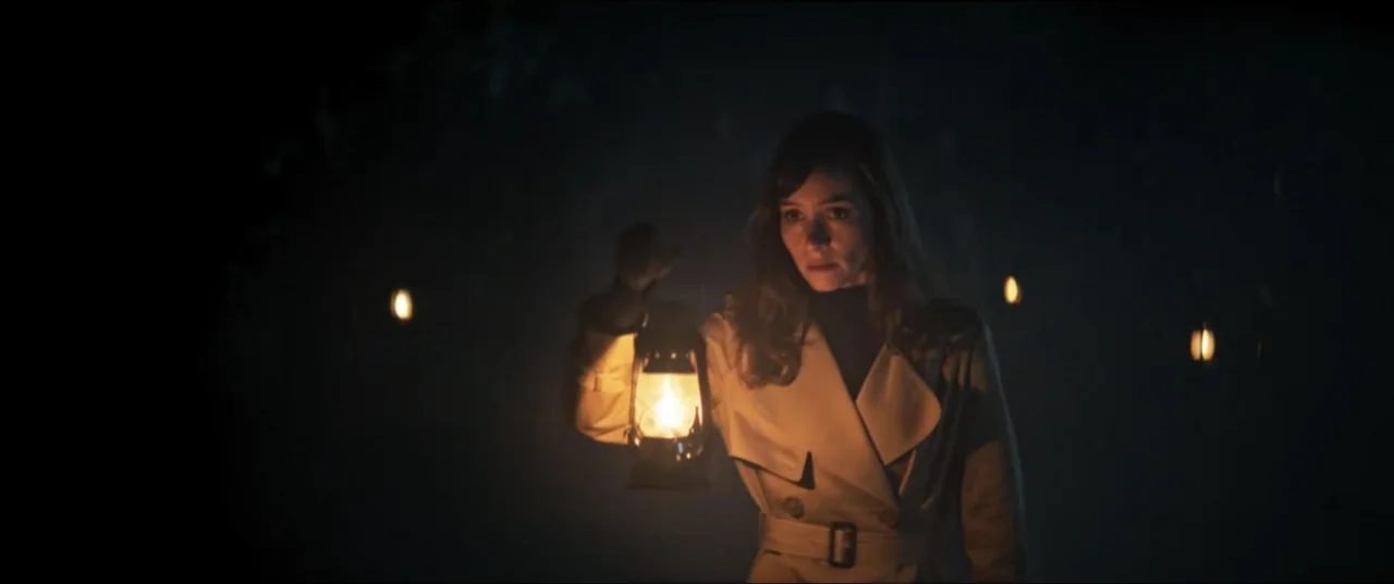 Offseason (2022) movie still: medium — A dark, dense outdoor environment is shrouded in deep shadows at night. A woman in a trench coat holds a…