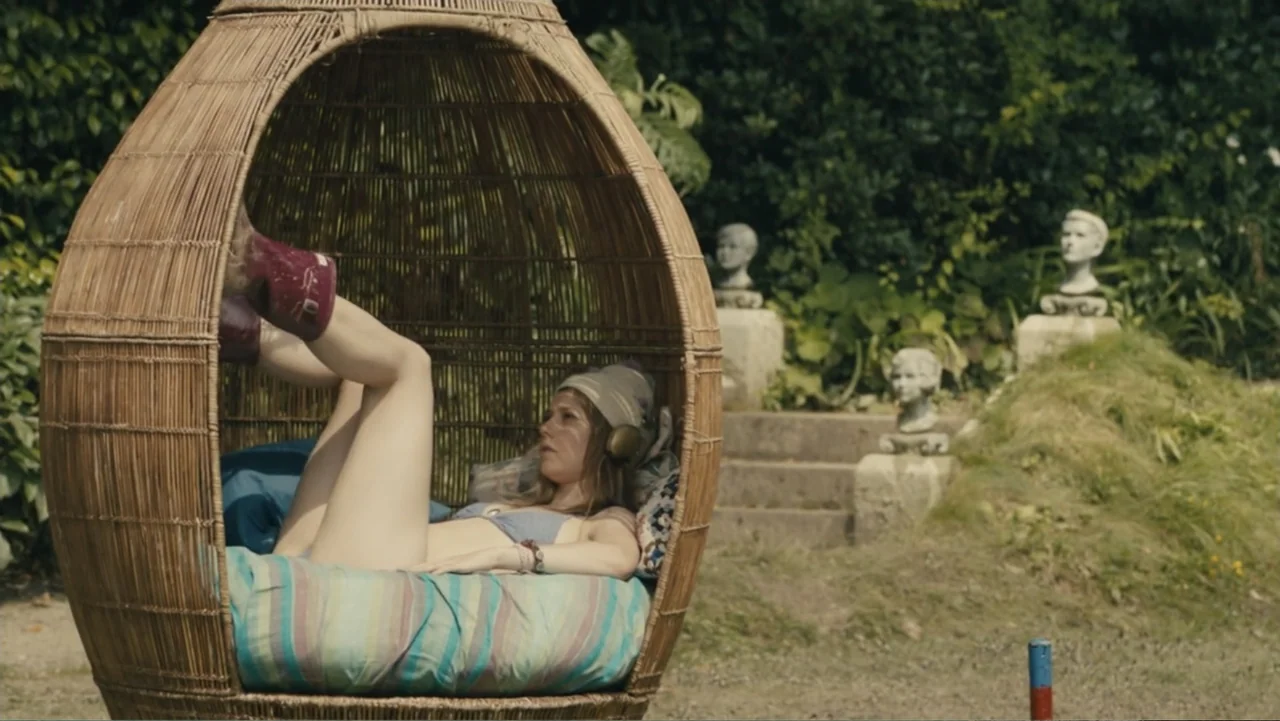 About Time (2013) movie still: medium — A wicker egg chair sits in a green backyard with stone sculptures in the background. A young woman reclines…