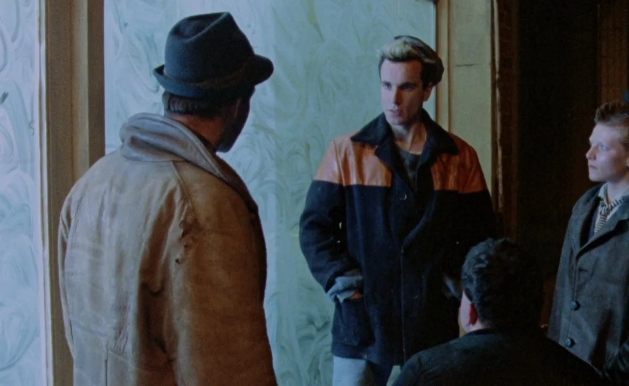 My Beautiful Laundrette (1985) movie still: medium — A sparse room features a large window with white swirling patterns on the glass. Four men stand…