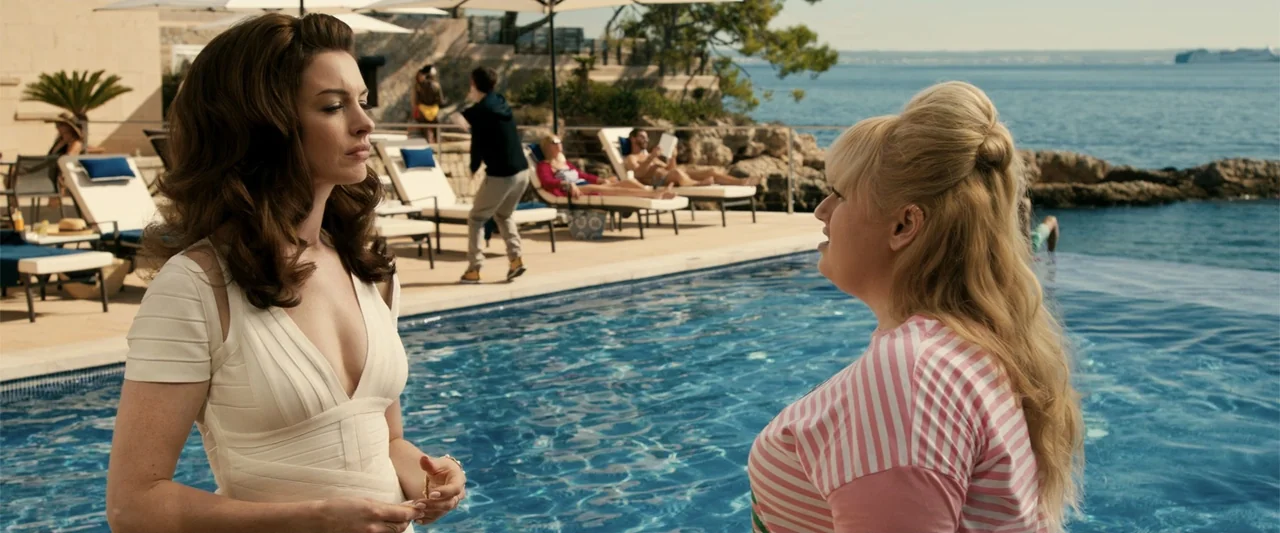 The Hustle (2019) movie still: medium — A sunny seaside infinity pool with lounge chairs and Mediterranean coastal rock formations. Two women face…