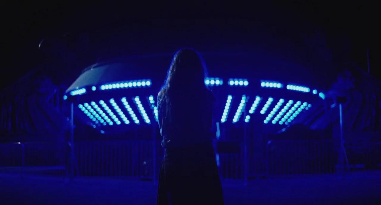 The Giant (2019) movie still: fullbody — A dark outdoor carnival at night with a large ride featuring glowing blue lights. A young person stands…
