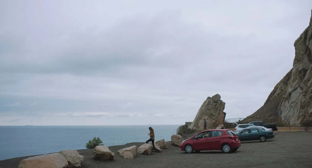 She Said (2022) movie still: wide — A gravel parking area sits atop a cliff overlooking a calm, vast ocean. A woman stands near the edge among large…
