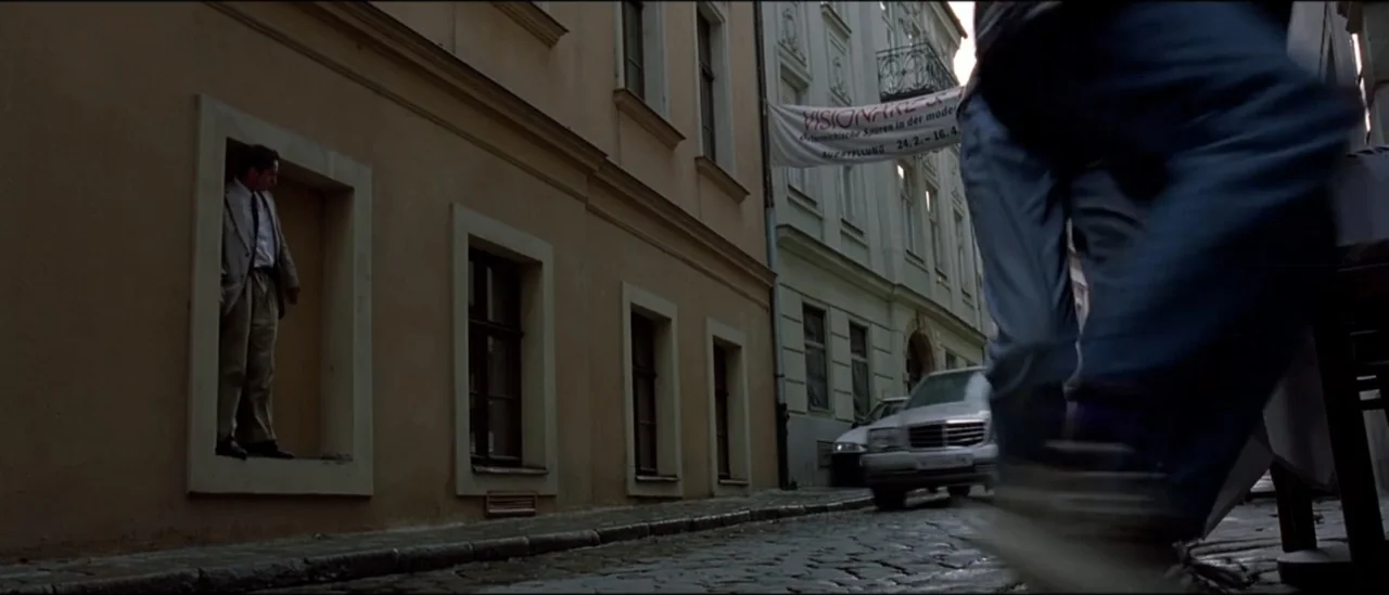 The Peacemaker (1997) movie still: wide — A European cobblestone street lined with beige buildings features a man in a window. A second person in…