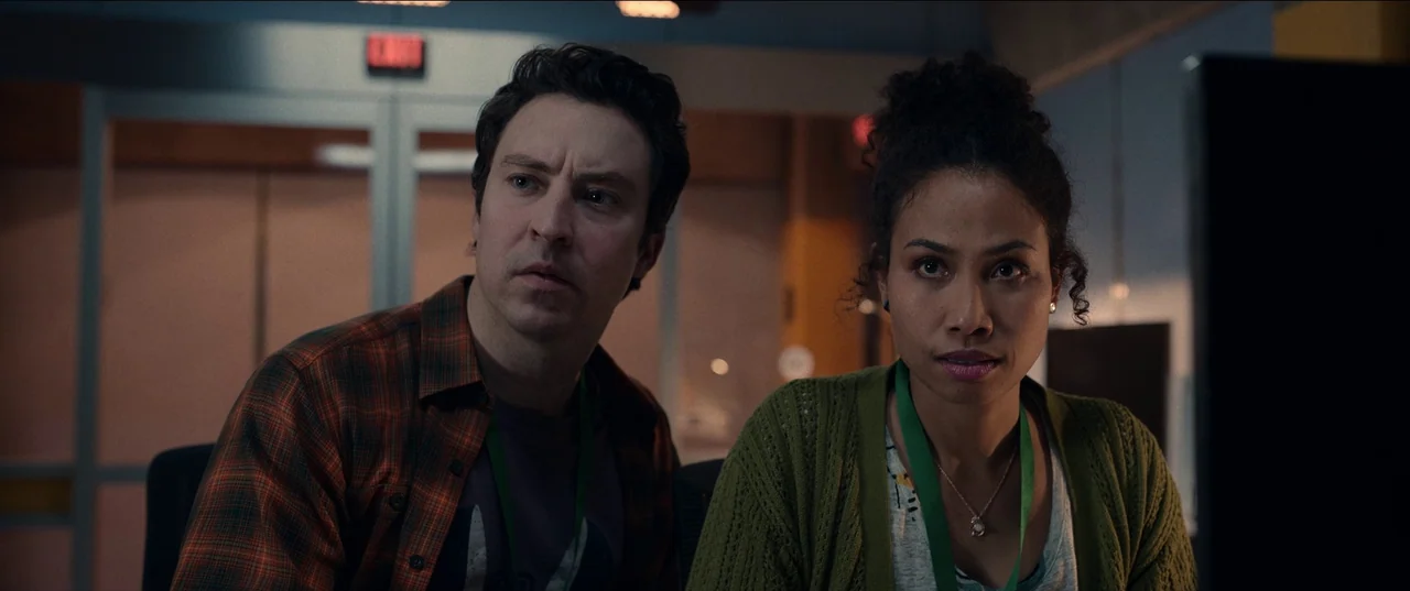 M3gan (2022) movie still: medium — A modern, dimly lit workspace featuring glass partitions and a glowing red exit sign. Two colleagues stand…