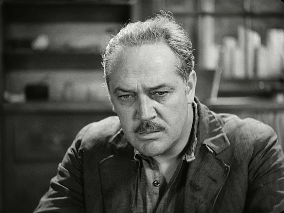 Modern Times (1936) movie still: closeup — A dimly lit indoor space with indistinct furniture and shelves in the background. A man sits in the…