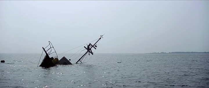 Jaws movie still: wide — An open expanse of grey sea surrounds a small sailboat sinking into the water. A lone person sits at the tip of the…