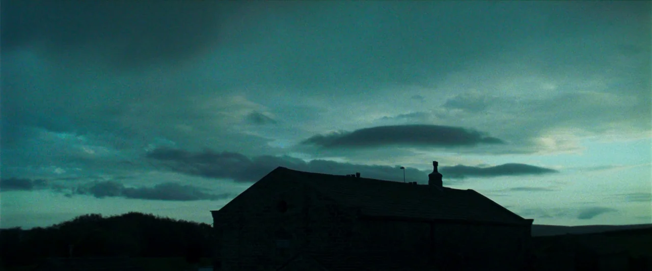 Starve Acre (2023) movie still: establishing — A stone farmhouse roofline stands silhouetted against a vast and cloudy blue twilight sky. The…