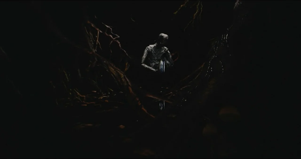 Eye for an Eye (2025) movie still: wide — A dark swamp area is filled with thick, twisted wooden tree roots. A pale humanoid figure stands still in…