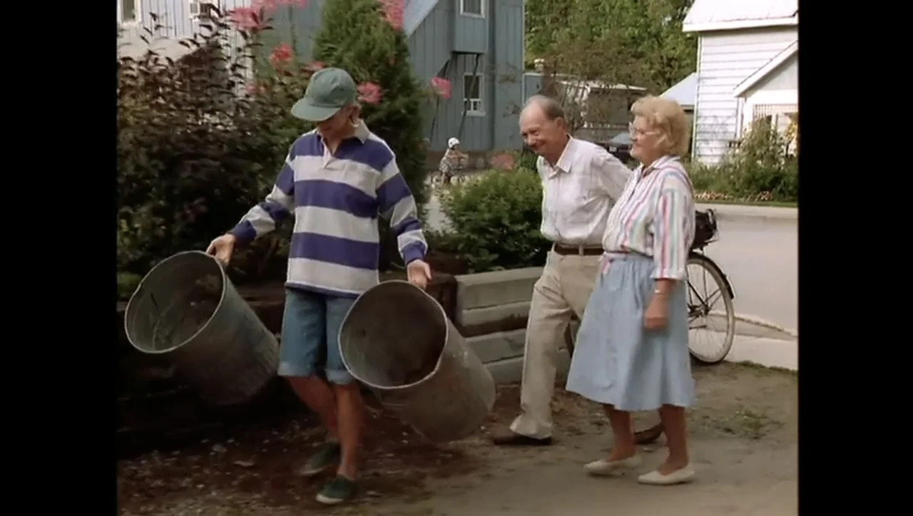 The Awakening (1995) movie still: fullbody — An exterior residential driveway features a person carrying two metal buckets past two elders. The…