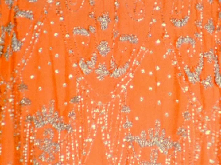 Puce Moment (1949) movie still: closeup — A close-up view shows orange fabric covered in intricate silver beadwork and embroidery. The pattern…