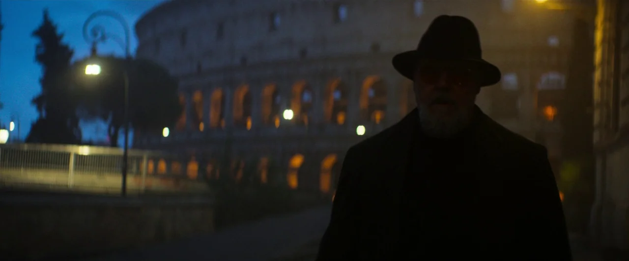 The Pope’s Exorcist (2023) movie still: medium — A dark, paved city street is illuminated by glowing streetlamps at night. A bearded man in a hat…