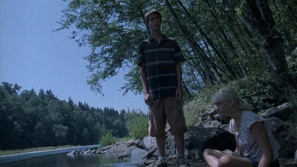 Mean Creek (2004) movie still: fullbody — A riverbank is lined with dense forest trees under a clear blue sky. A boy stands on the edge of the water…