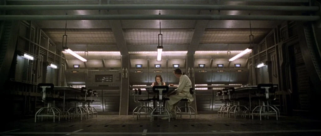 Alien: Resurrection (1997) movie still: wide — A spacious, metallic industrial mess hall features rows of tables and modular wall panels. Two…