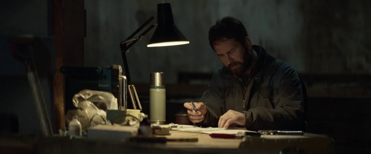 Babel (2006) movie still: medium — A dimly lit industrial workspace with a cluttered wooden desk and dark concrete walls. A man in a work jacket…